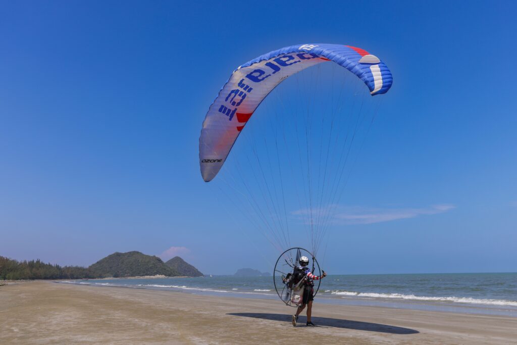 A paraglider with motorized equipment prepares for takeoff on a scenic beach under a clear blue sky.