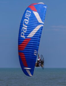 A paramotor aerodynamically gliding over ocean waters with a prominent blue and red parachute against a clear sky.